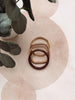 Three small interlocking ring-shaped accessories in gold, rose gold, and dark burgundy, arranged on a pale pink textured surface with decorative green leaves in the corner.