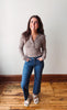 A woman stands in a room with a white wall and wooden floor, wearing a brown and beige striped long-sleeve henley top, blue jeans, and white slide sandals.