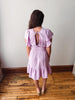 Woman wearing a light purple dress with ruffled sleeves and open back, standing on a wooden floor.