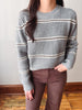 A person wearing a gray ribbed knit sweater with thin horizontal black and white stripes, paired with brown pants, standing indoors against a plain wall and wood floor.