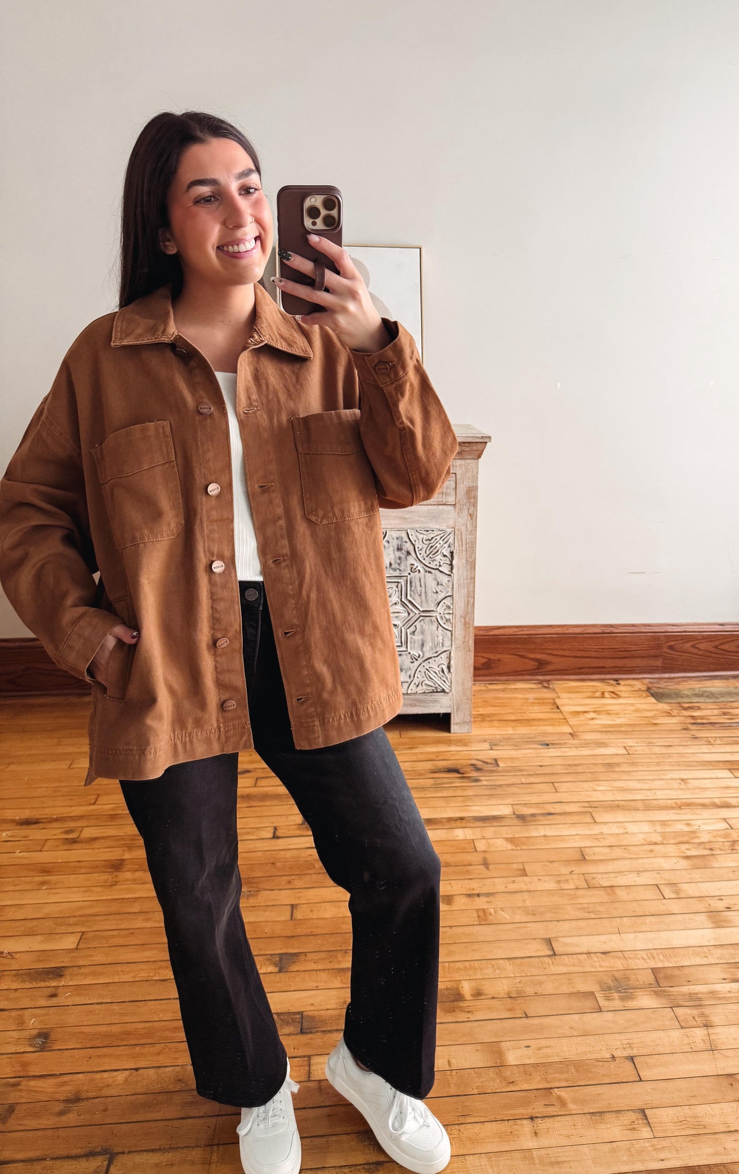 A woman takes a mirror selfie wearing a brown corduroy button-up shirt jacket over a white t-shirt, black wide-leg pants, and white sneakers in a room with wooden floors and a white wall.