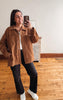 A woman takes a mirror selfie wearing a brown corduroy button-up shirt jacket over a white t-shirt, black wide-leg pants, and white sneakers in a room with wooden floors and a white wall.