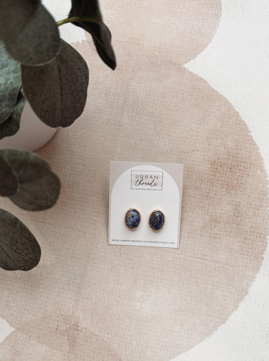 A pair of oval stud earrings with dark blue and white marbled inlay and gold-tone edges, displayed on a white backing card labeled Urban Jewelry, styled on a textured beige surface with a plant leaf in the corner.
