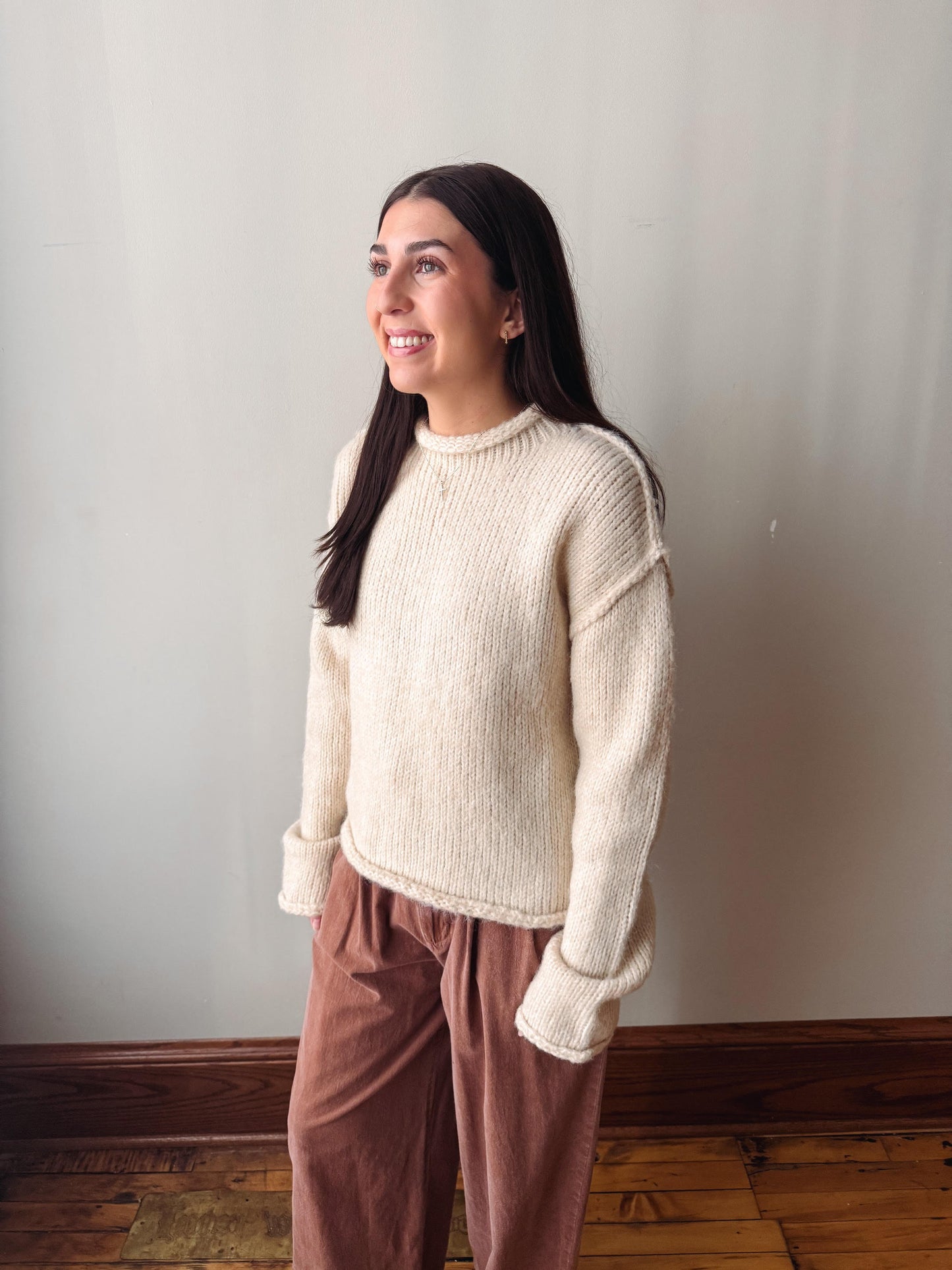 A woman wearing a cream ribbed knit mock neck sweater and dusty rose corduroy pants, standing against a plain light gray wall on a wooden floor.
