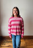 A smiling woman with shoulder-length brown hair stands indoors wearing a pink and light pink horizontally striped knit sweater and blue jeans against a plain off-white wall and wooden floor.