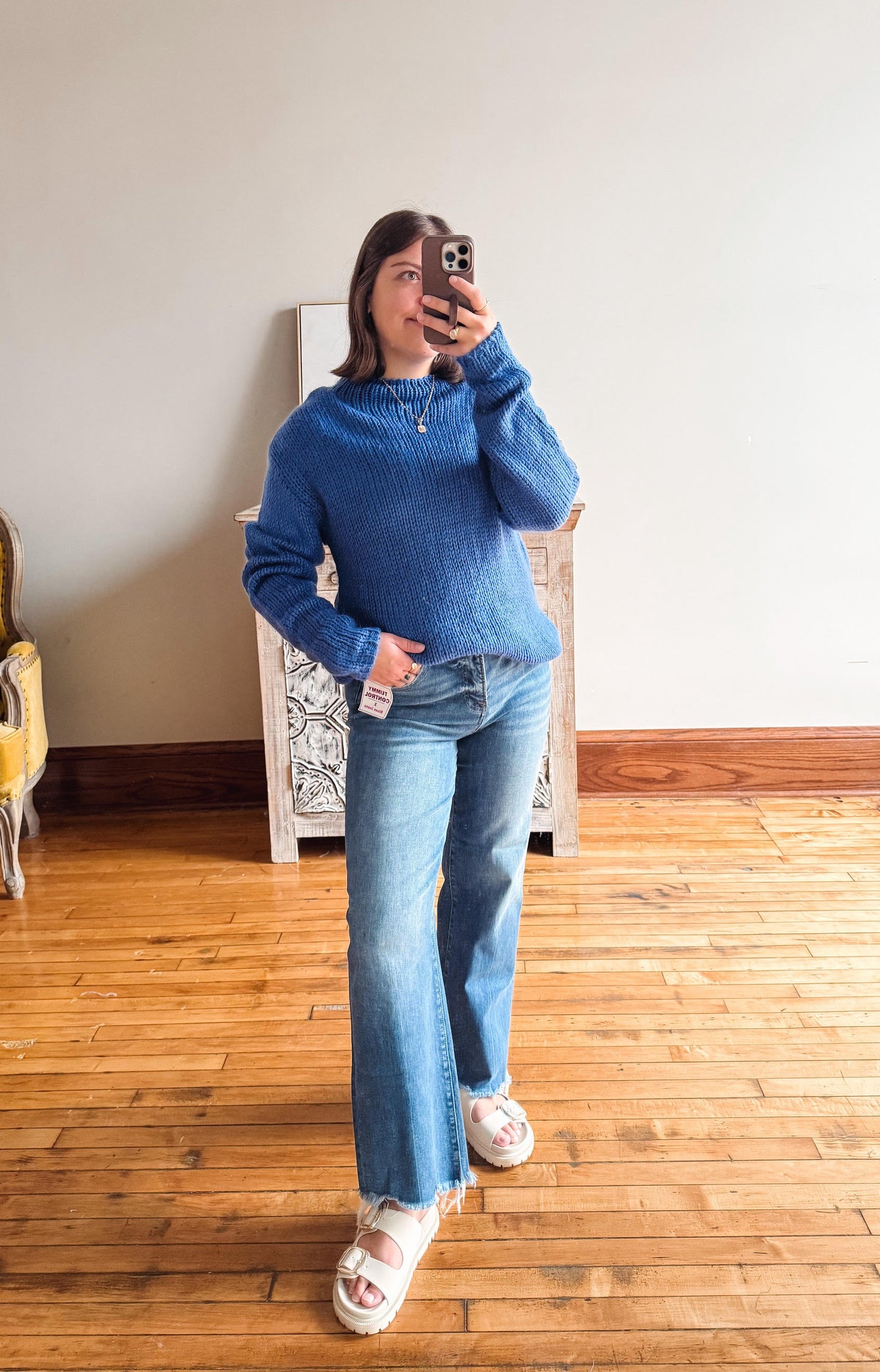 A woman takes a mirror selfie wearing a blue ribbed knit sweater and light-wash flare jeans with white slide sandals, standing in a room with hardwood floors and a white wall.