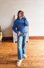 A woman takes a mirror selfie wearing a blue ribbed knit sweater and light-wash flare jeans with white slide sandals, standing in a room with hardwood floors and a white wall.