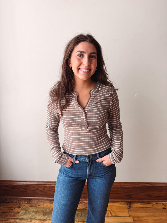 A smiling woman with long brown hair stands indoors wearing a brown and white striped long-sleeve henley top and blue jeans, with her hands in her pockets, against a plain off-white wall and wooden floor.