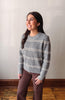 A woman wearing a gray ribbed knit sweater with horizontal cream stripes and brown pants, standing in a room with a light gray wall and wooden floor.