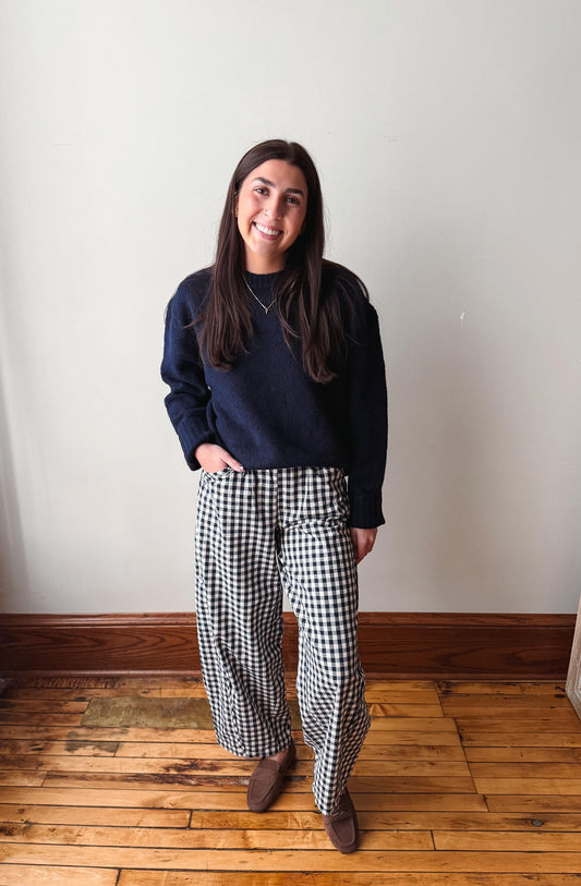 A woman stands indoors against a plain off-white wall on a wooden floor, wearing a dark navy crewneck sweatshirt and black-and-white gingham wide-leg pants with brown shoes.