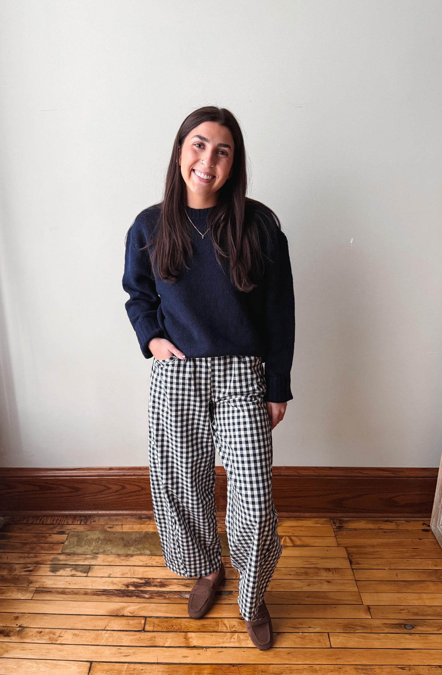 A woman stands indoors against a plain off-white wall on a wooden floor, wearing a dark navy crewneck sweatshirt and black-and-white gingham wide-leg pants with brown shoes.