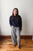 A woman stands indoors against a plain off-white wall on a wooden floor, wearing a dark navy crewneck sweatshirt and black-and-white gingham wide-leg pants with brown shoes.