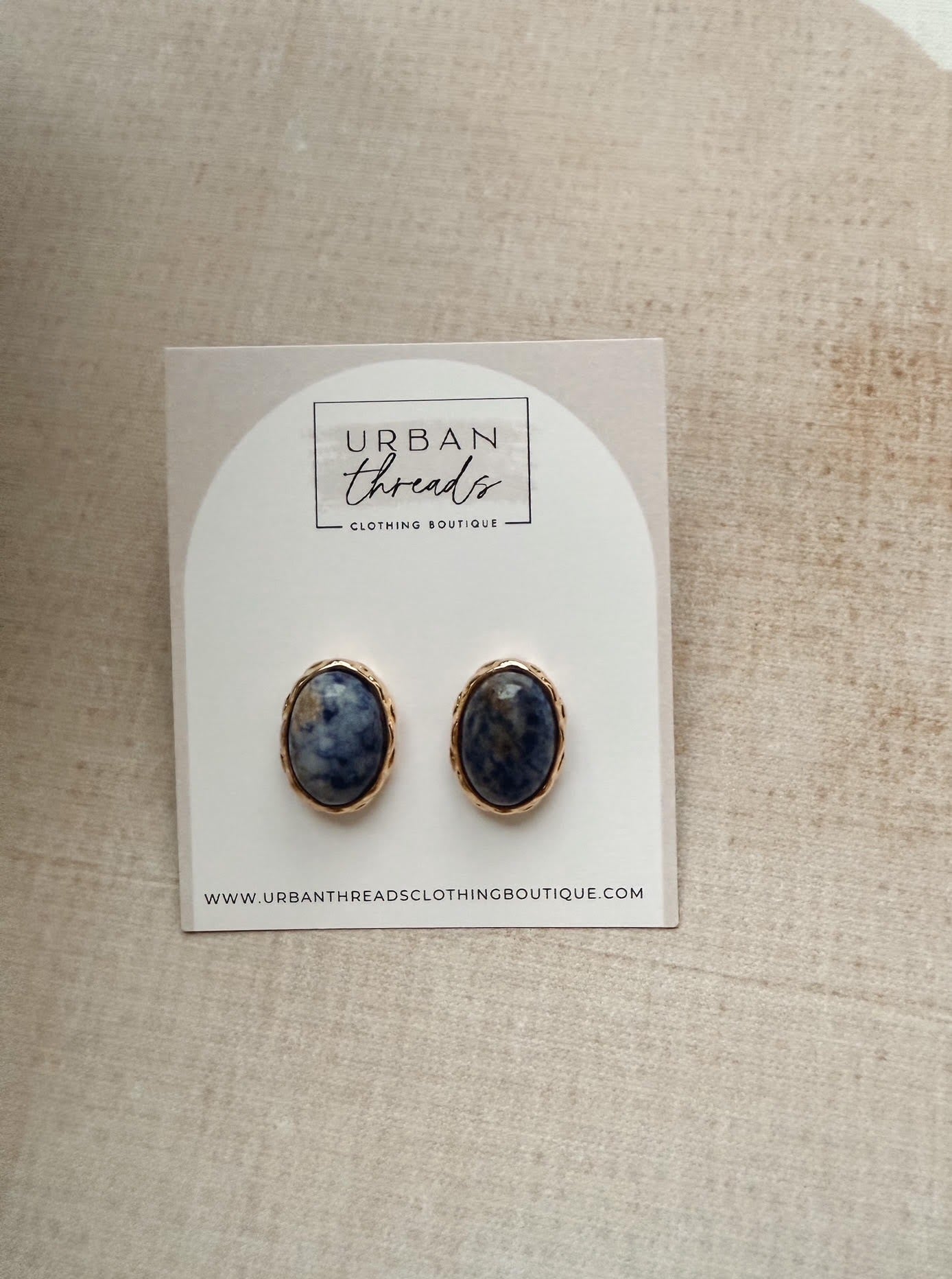 A pair of oval stud earrings with dark blue and white marbled stones in gold bezel settings, displayed on a white arched backing card labeled Urban Threads, set against a beige linen background.