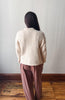 A woman viewed from behind wearing a cream ribbed knit cropped cardigan and dusty rose wide-leg trousers, standing on a wooden floor against a plain light gray wall.
