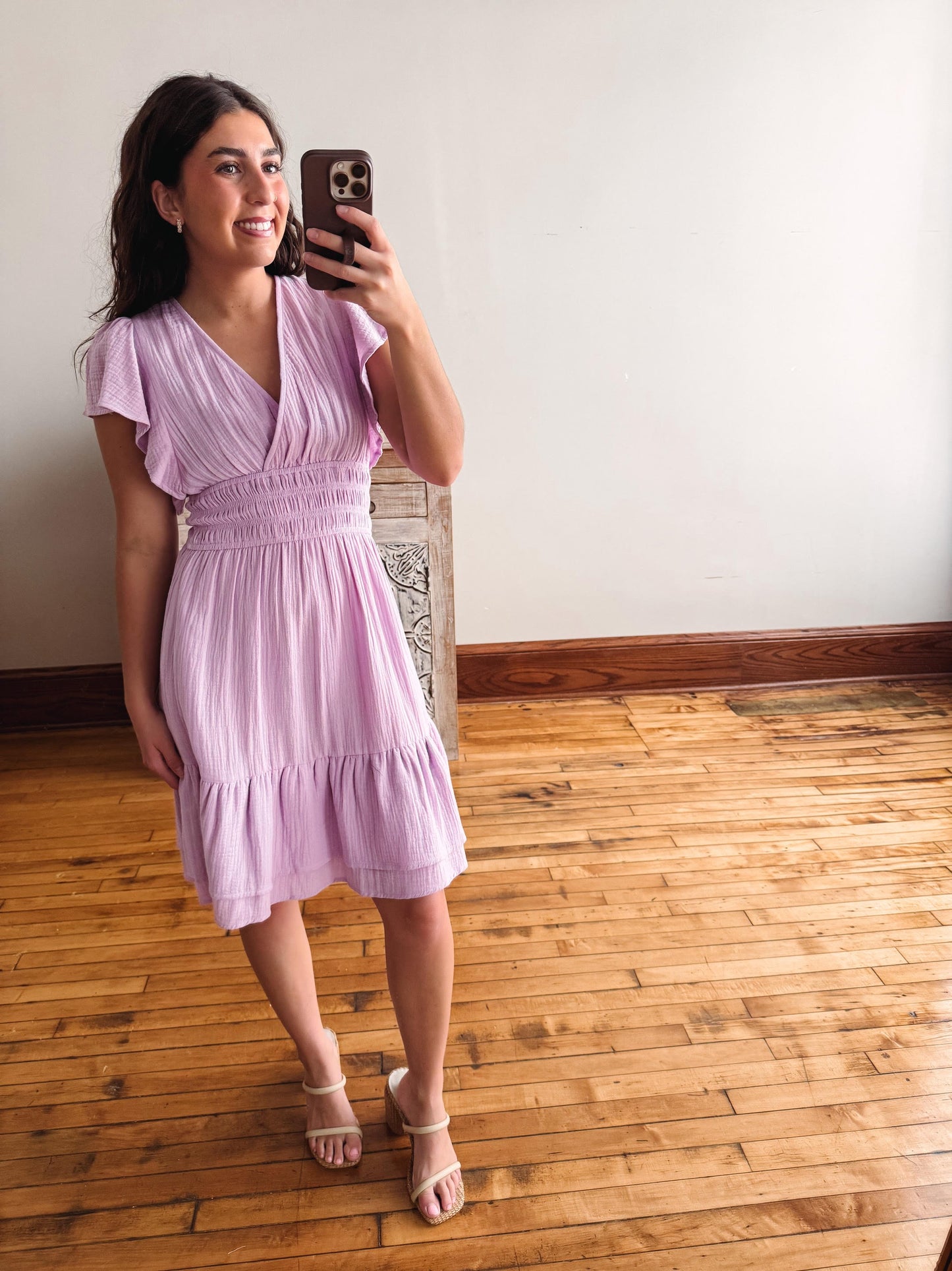 Woman taking a mirror selfie wearing a light purple dress on a wooden floor.