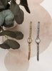 Two slim analog watches with oval crystal cases are displayed vertically on a light beige surface with soft pink abstract shapes. One watch has a gold-tone chain and case, and the other has a silver-tone chain and case.