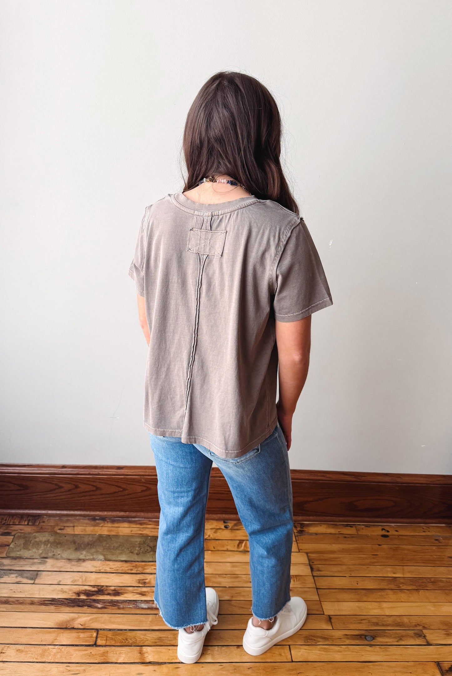 Person wearing a beige t-shirt and blue jeans standing on a wooden floor with a white wall background