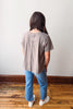 Person wearing a beige t-shirt and blue jeans standing on a wooden floor with a white wall background