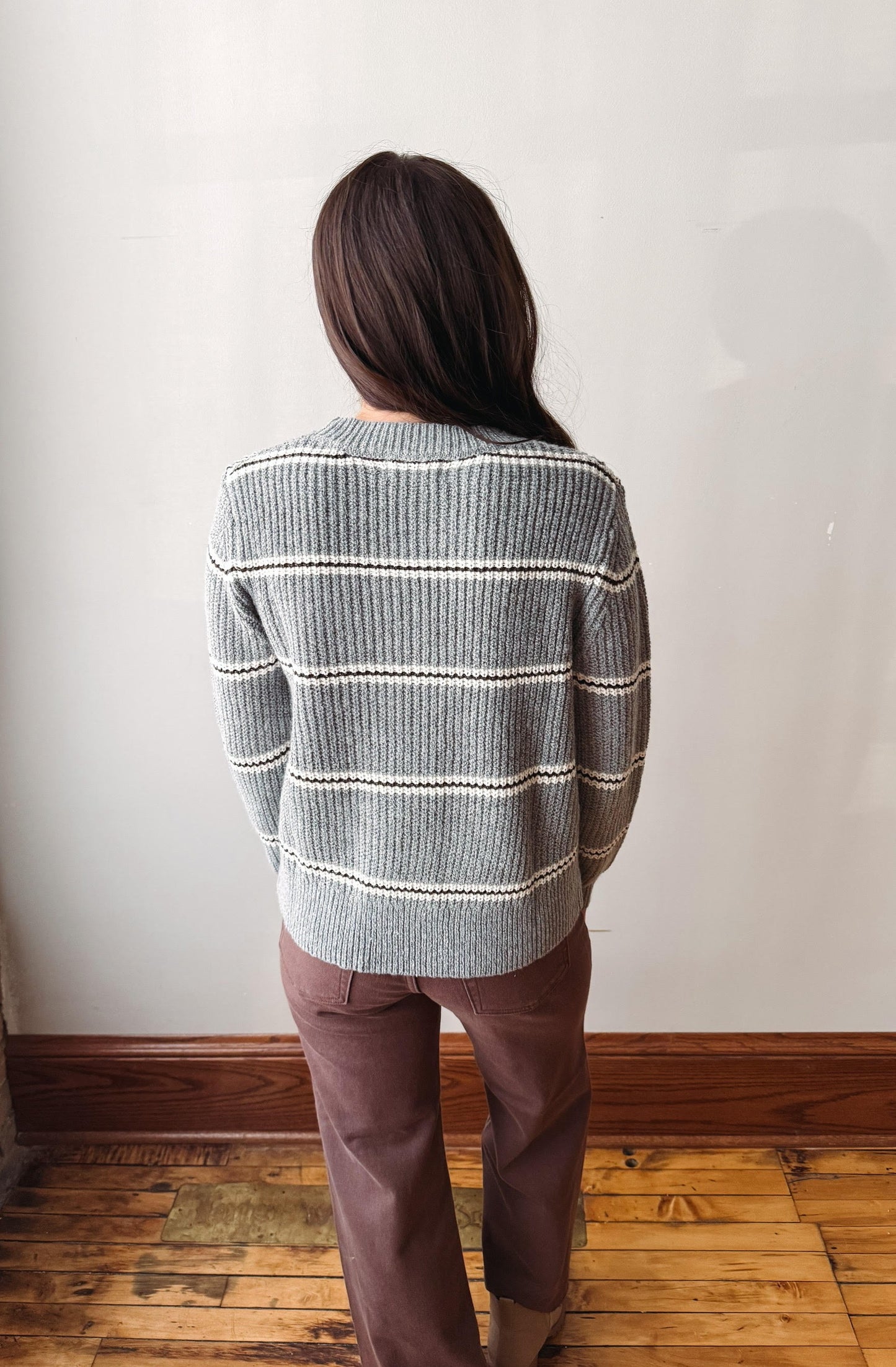 A woman viewed from behind wearing a gray ribbed knit sweater with thin white horizontal stripes and brown pants, standing on a wooden floor against a plain off-white wall.