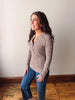A woman wearing a brown and white striped long-sleeve henley top and blue jeans, standing in a room with a light beige wall and wooden floor.