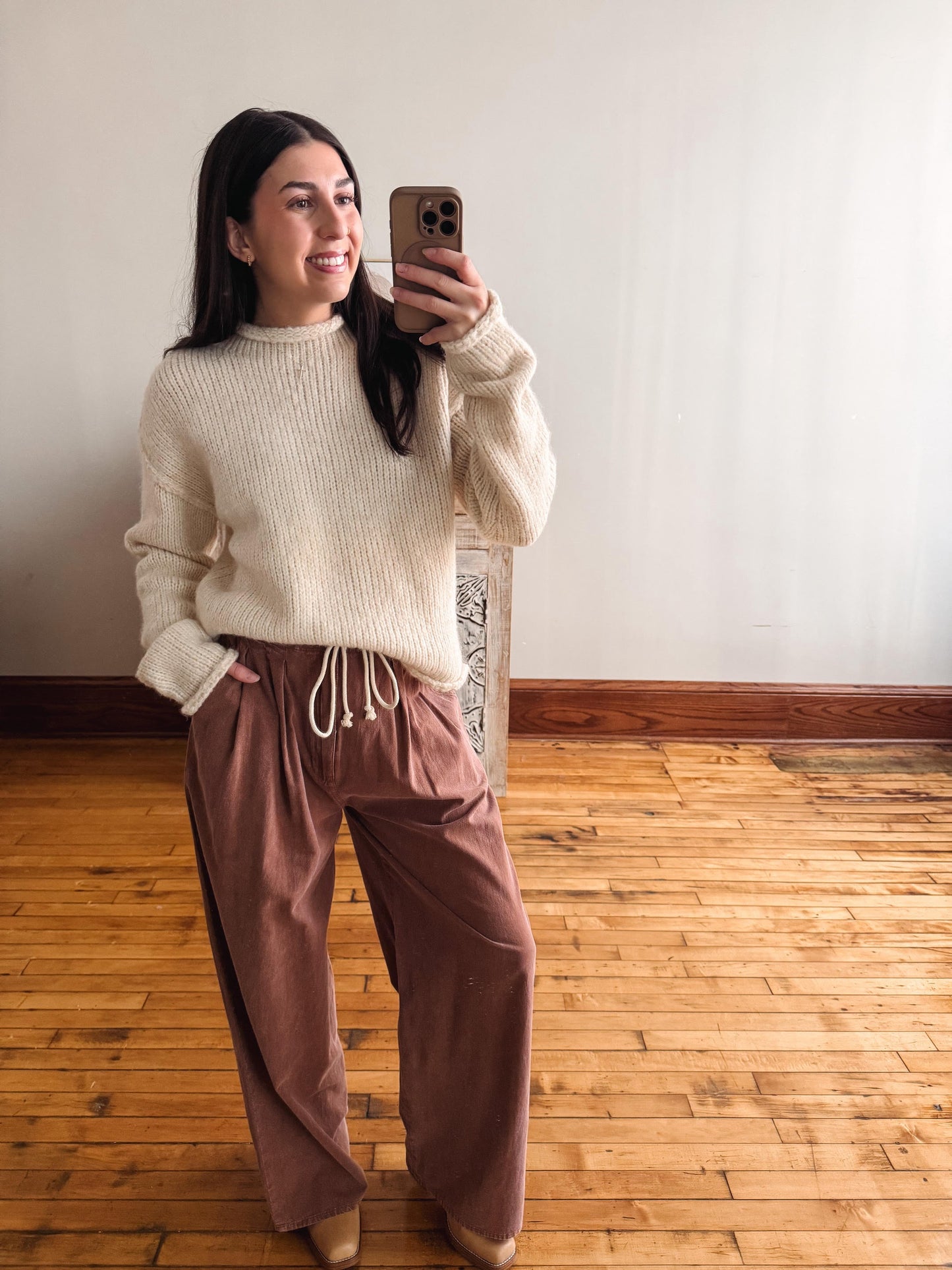 A woman taking a mirror selfie in a room with light-colored walls and a wooden floor, wearing a cream ribbed knit sweater and dusty rose wide-leg pants.