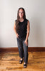 Woman wearing a black sleeveless top and dark jeans standing on a wooden floor with a white wall background.