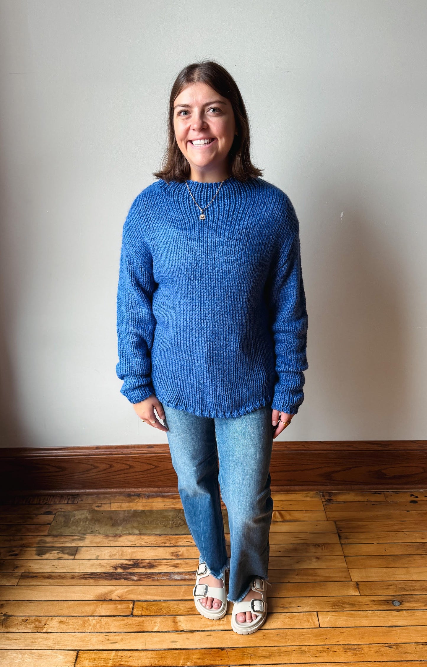 A woman wearing a blue textured knit sweater, blue jeans, and white slide sandals, standing on a wooden floor against a plain off-white wall.