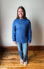 A woman wearing a blue textured knit sweater, blue jeans, and white slide sandals, standing on a wooden floor against a plain off-white wall.