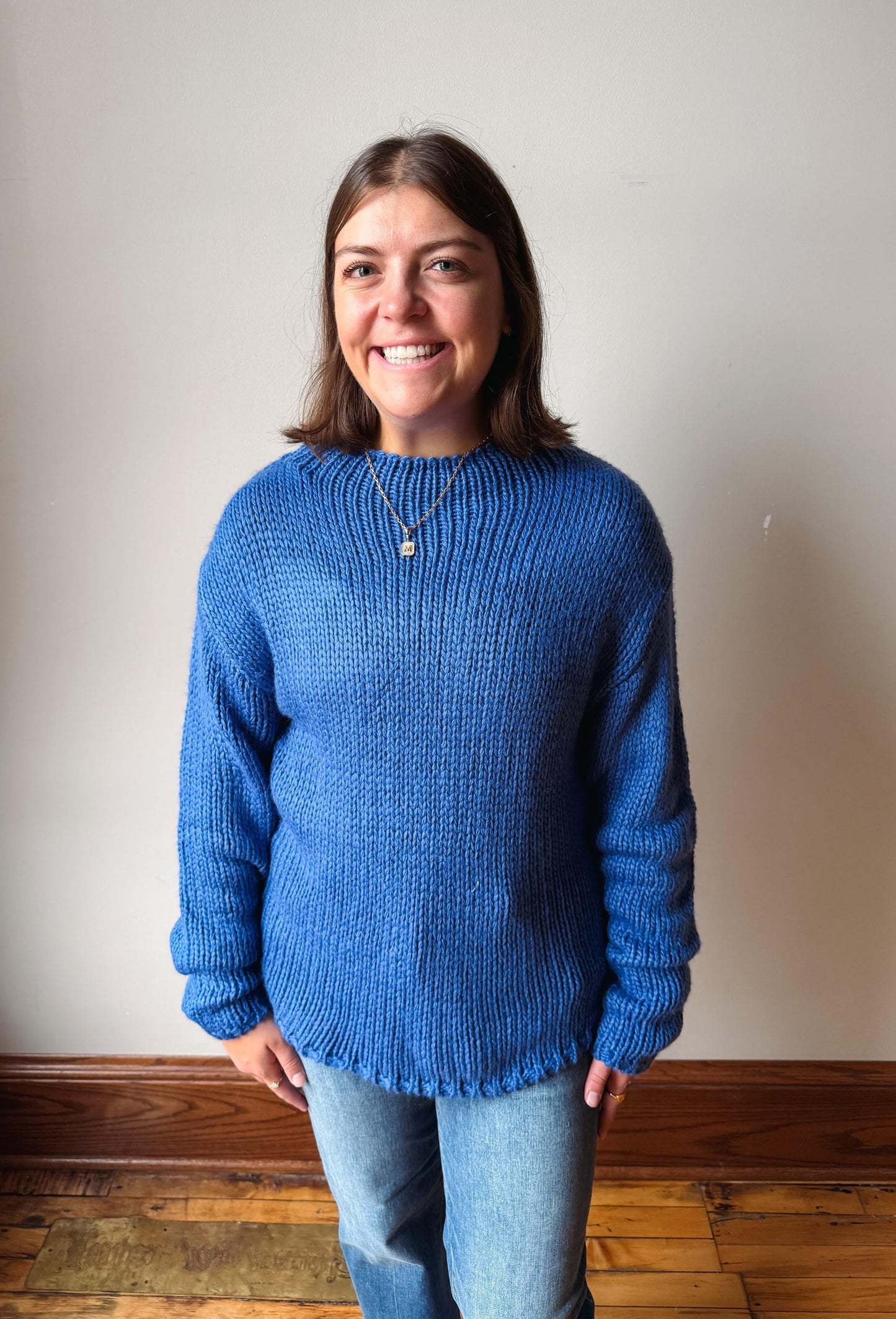 A smiling woman with shoulder-length brown hair stands indoors wearing a chunky knit blue sweater and light blue jeans against a plain off-white wall with a wooden baseboard and floor.