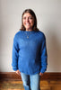 A smiling woman with shoulder-length brown hair stands indoors wearing a chunky knit blue sweater and light blue jeans against a plain off-white wall with a wooden baseboard and floor.