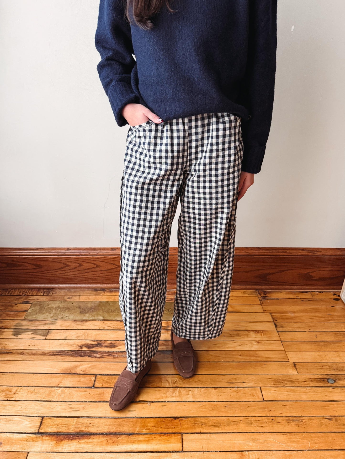 A person wearing black and white gingham wide-leg trousers, a navy blue sweater, and brown suede loafers, standing on a wooden floor against a white wall.