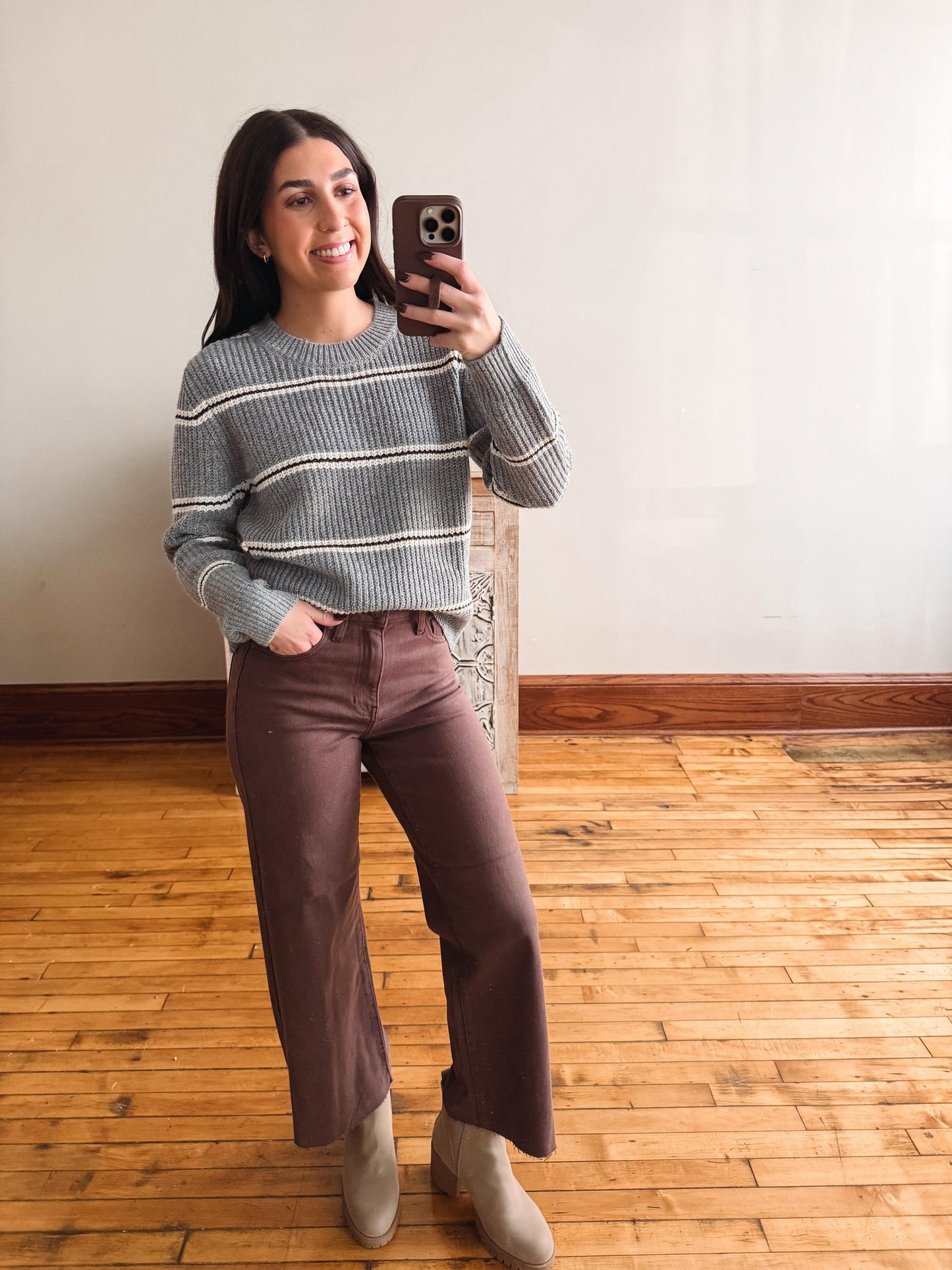 A woman takes a mirror selfie in a room with a light-colored wall and wooden floor, wearing a gray and white striped knit sweater, brown wide-leg pants, and beige ankle boots.