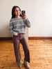 A woman takes a mirror selfie in a room with a light-colored wall and wooden floor, wearing a gray and white striped knit sweater, brown wide-leg pants, and beige ankle boots.