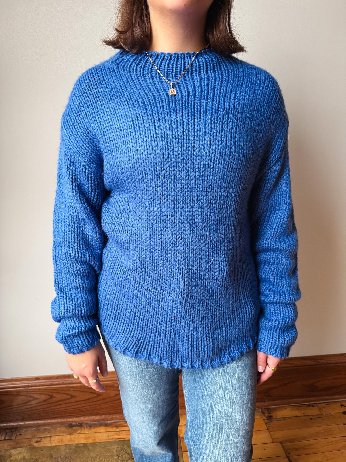 A person wearing a chunky blue knit sweater with a crew neck and ribbed texture, paired with light blue jeans, standing against a plain beige wall with a wooden floor.