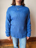 A person wearing a chunky blue knit sweater with a crew neck and ribbed texture, paired with light blue jeans, standing against a plain beige wall with a wooden floor.