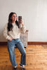 A woman takes a mirror selfie in a room with a light-colored wall, wooden floor, and a small cabinet. She is wearing a cream-colored textured knit sweater, blue wide-leg jeans, and brown sneakers.