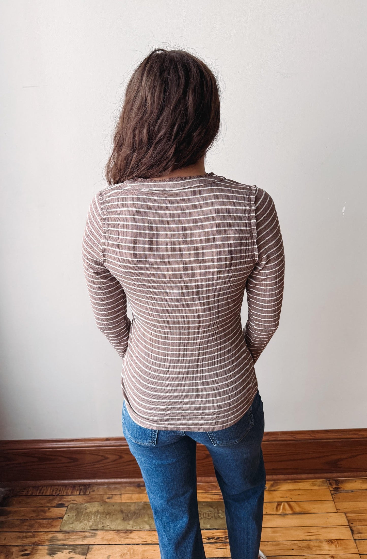 A woman viewed from behind wearing a brown and white horizontally striped long-sleeve top and blue jeans, standing in a room with a white wall and wooden floor.