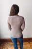 A woman viewed from behind wearing a brown and white horizontally striped long-sleeve top and blue jeans, standing in a room with a white wall and wooden floor.