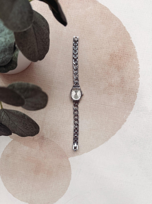 A silver-toned women's watch with a textured link bracelet and a round silver dial, displayed vertically on a beige fabric surface with watercolor-style circles and dark green leaves in the corner.