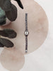 A silver-toned women's watch with a textured link bracelet and a round silver dial, displayed vertically on a beige fabric surface with watercolor-style circles and dark green leaves in the corner.