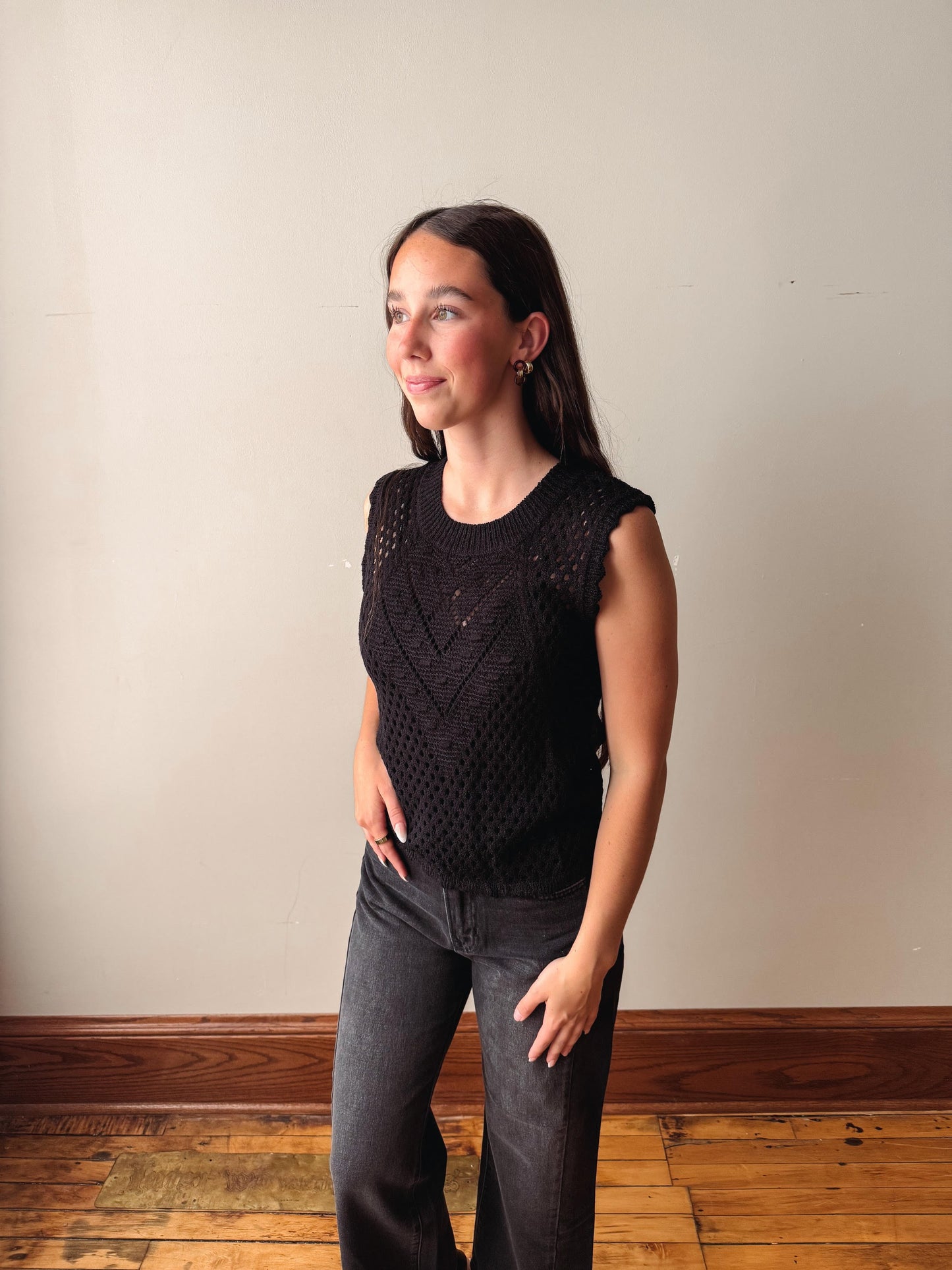 Woman wearing a black sleeveless knitted top and dark jeans against a plain wall.