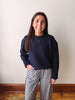 A woman smiling and posing indoors, wearing a dark navy blue crewneck sweater and black-and-white gingham pants, standing against a plain off-white wall with wooden baseboards and floorboards visible.