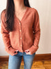 A person wearing a rust-colored knit cardigan sweater with buttons, paired with blue jeans, standing indoors against a plain wall.