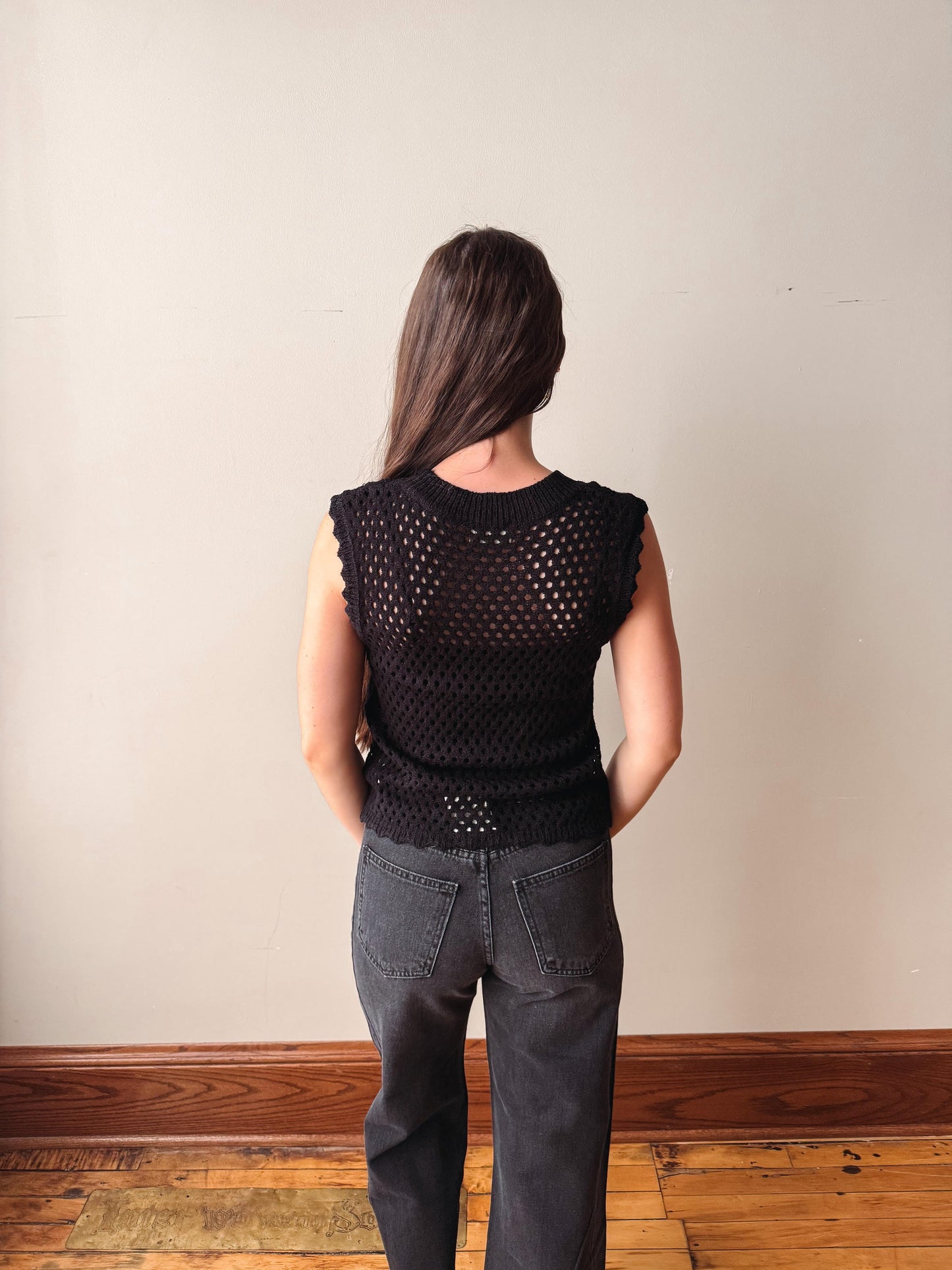 Person wearing a black sleeveless top and gray pants against a plain wall.