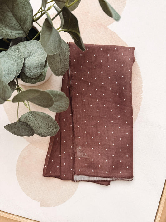 A folded brown fabric scarf with small white polka dots, styled on a light beige background with a green eucalyptus plant.