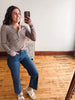 A woman takes a mirror selfie in a room with hardwood floors and a white wall, wearing a brown and beige horizontally striped long-sleeve henley top, blue jeans, and white slide sandals.