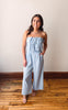 Woman wearing a light blue strapless jumpsuit standing on a wooden floor with a plain wall background
