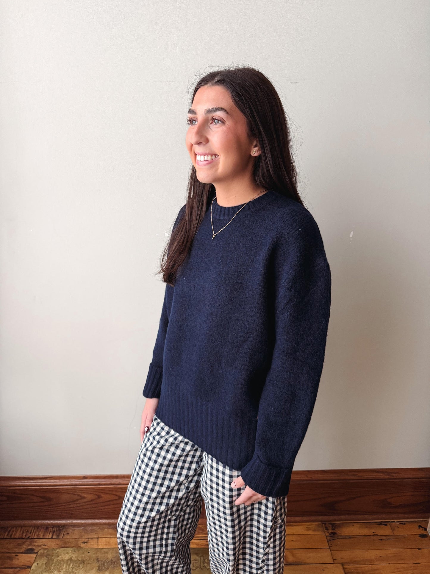 A woman wearing a dark navy blue knit sweater and black-and-white gingham pants, standing in a room with a beige wall and wooden floor.
