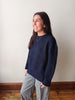 A woman wearing a dark navy blue knit sweater and black-and-white gingham pants, standing in a room with a beige wall and wooden floor.