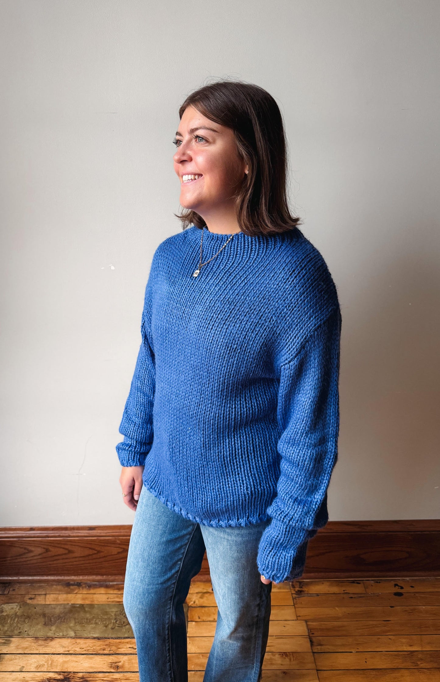 A woman wearing a blue chunky knit sweater and blue jeans, standing in a room with a light gray wall and wooden floor.